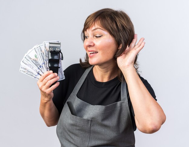 pleased adult female barber in uniform holding hair clipper with money and keeping hand close to ear trying to hear isolated on white wall with copy space