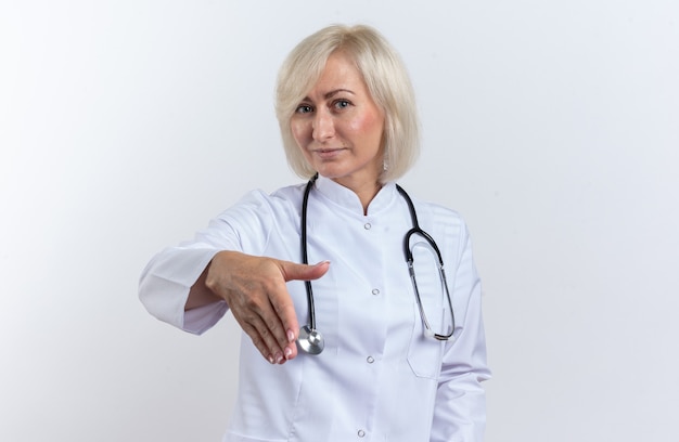 Free photo pleased adult female doctor in medical robe with stethoscope holding out her hand isolated on white wall with copy space