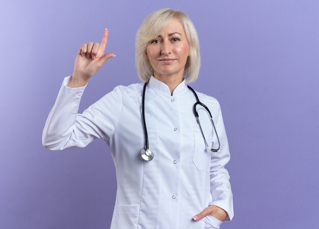 Довольная взрослая женщина-врач в медицинском халате со стетоскопом, направленным вверх изолирована на фиолетовой стене с копией пространства