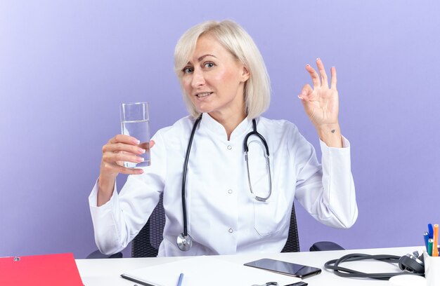 聴診器が机に座って、水のガラスを保持し、コピースペースのある紫色の壁に隔離されたokサインを身振りで示す医療ローブの大人の女性医師を喜ばせます