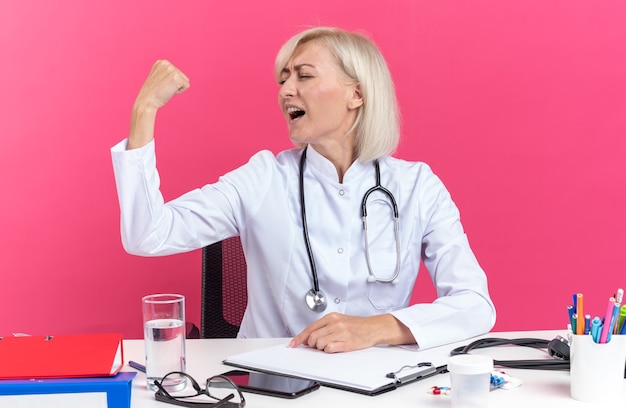 довольная взрослая женщина-врач в медицинском халате со стетоскопом сидит за столом с офисными инструментами, напрягая бицепсы, изолированные на розовой стене с копией пространства