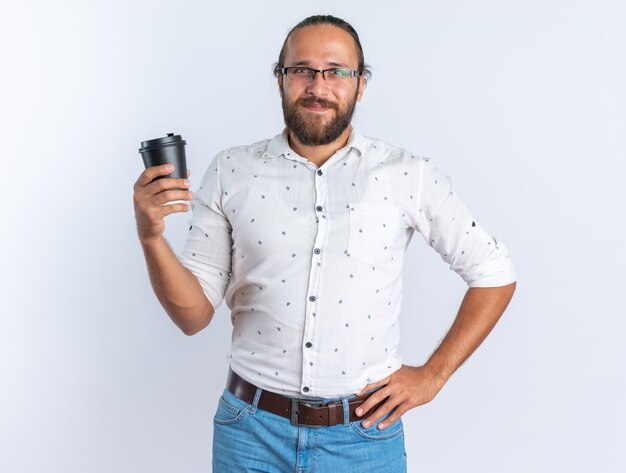 Pleased adult handsome man wearing glasses keeping hand on waist holding plastic coffee cup looking at camera isolated on white wall