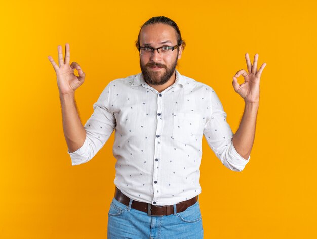 Pleased adult handsome man wearing glasses looking at camera doing ok sign isolated on orange wall