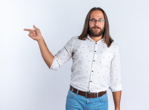 Pleased adult handsome man wearing glasses looking at camera pointing at side isolated on white wall