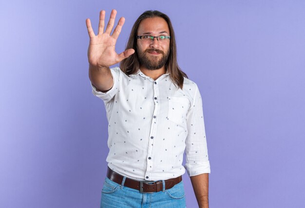 Pleased adult handsome man wearing glasses looking at camera showing five with hand isolated on purple wall with copy space