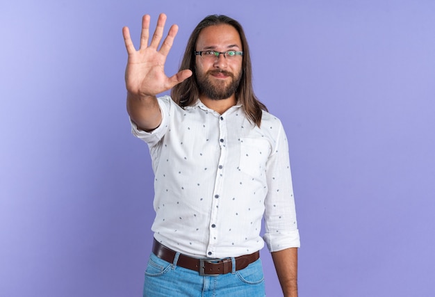 Pleased adult handsome man wearing glasses looking at camera showing five with hand isolated on purple wall with copy space