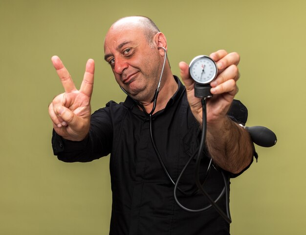 pleased adult ill caucasian man holding sphygmomanometer and gesturing victory sign isolated on olive green wall with copy space