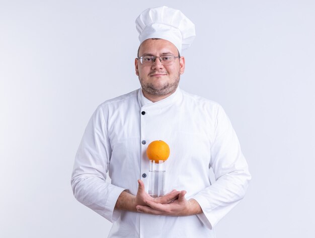 pleased adult male cook wearing chef uniform and glasses holding glass of water with orange on it looking at front isolated on white wall