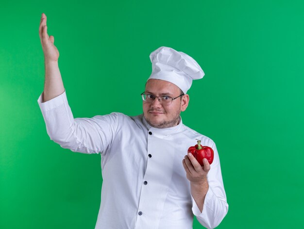 요리사 유니폼을 입고 안경을 쓴 성인 남성 요리사는 녹색 벽에 격리된 손을 위로 올리는 쪽을 바라보는 후추를 들고 기뻐했습니다.