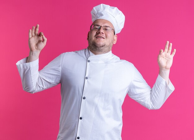 pleased adult male cook wearing chef uniform and glasses looking at front doing ok sign isolated on pink wall