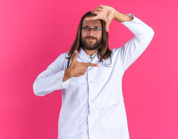 Pleased adult male doctor wearing medical robe and stethoscope with glasses looking at camera doing frame gesture isolated on pink wall