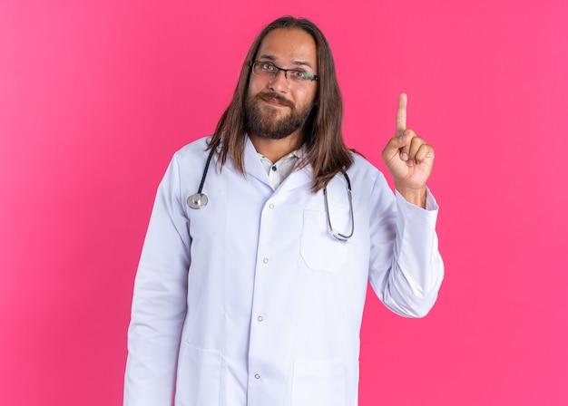 Pleased adult male doctor wearing medical robe and stethoscope with glasses looking at camera pointing up isolated on pink wall