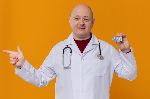 Pleased adult man in doctor uniform with stethoscope holding medicine blister pack and pointing at side 