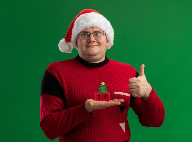 Pleased adult man wearing glasses and santa hat holding christmas tree toy with date  looking at side showing thumb up isolated on green background