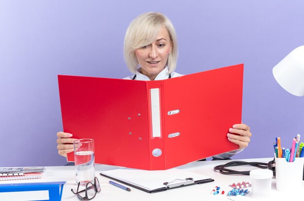 Pleased adult slavic female doctor in medical robe with stethoscope sitting at desk with office tools holding and looking at file folder isolated on purple wall with copy space