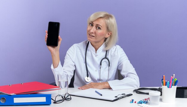 Pleased adult slavic female doctor in medical robe with stethoscope sitting at desk with office tools holding phone isolated on purple background with copy space