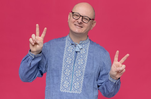 Pleased adult slavic man in blue shirt wearing optical glasses gesturing victory sign 