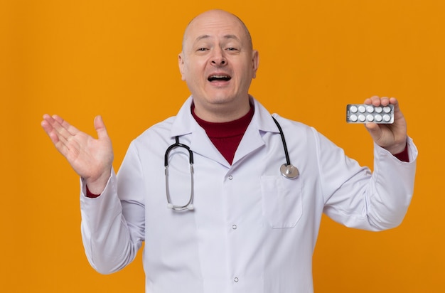 Pleased adult slavic man in doctor uniform with stethoscope holding medicine blister pack and keeping his hand open