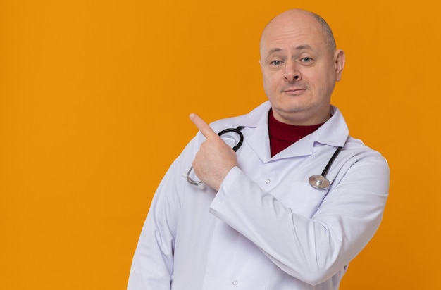 Pleased adult slavic man in doctor uniform with stethoscope pointing at side 