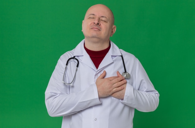 Pleased adult slavic man in doctor uniform with stethoscope putting hands on his chest 