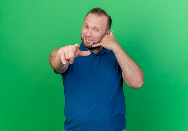 Pleased adult slavic man doing call gesture and pointing isolated on green wall with copy space