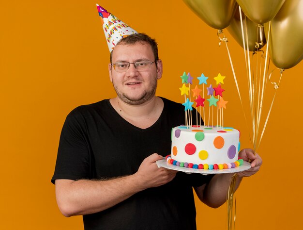 誕生日の帽子をかぶった光学眼鏡をかけた、満足している大人のスラブ人が、カメラを見てヘリウム風船と誕生日ケーキを持っている