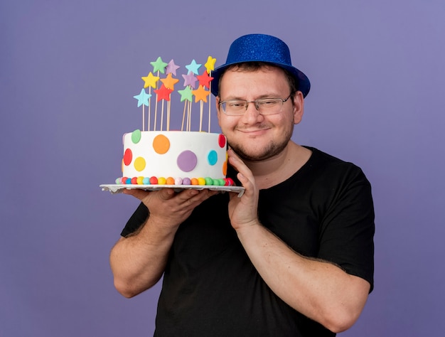 Pleased adult slavic man in optical glasses wearing blue party hat holds birthday cake 