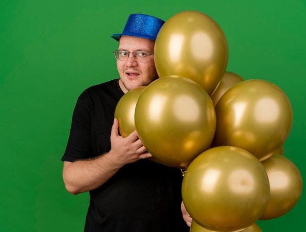 Pleased adult slavic man in optical glasses wearing blue party hat holds helium balloons 