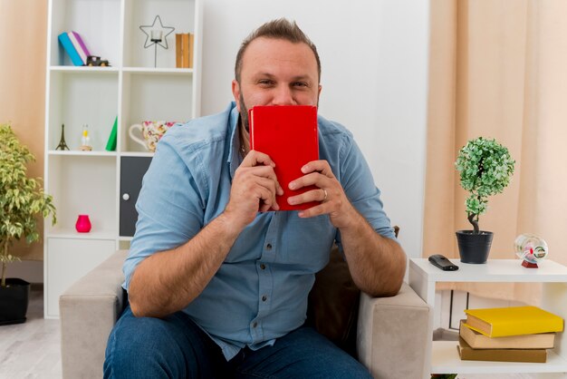 Довольный взрослый славянский мужчина сидит на кресле, держа книгу перед ртом в гостиной