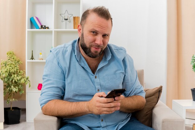 Pleased adult slavic man sits on armchair holding phone looking at camera inside the living room