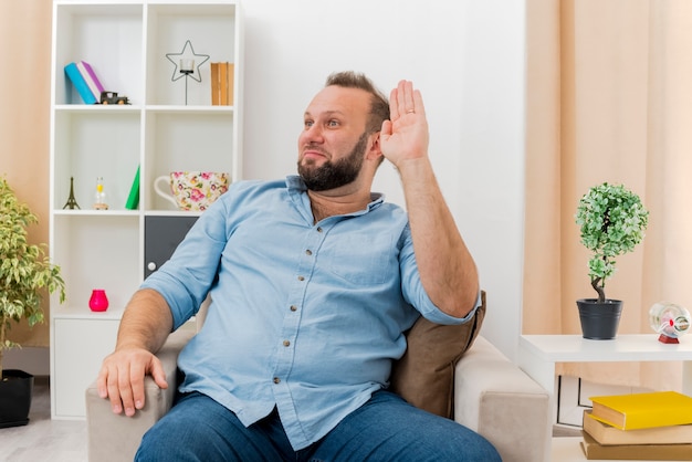 Pleased adult slavic man sits on armchair with raised hand looking at side inside the living room