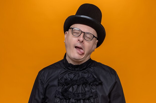 Pleased adult slavic man with top hat and optical glasses in black gothic shirt stucks out tongue standing with closed eyes