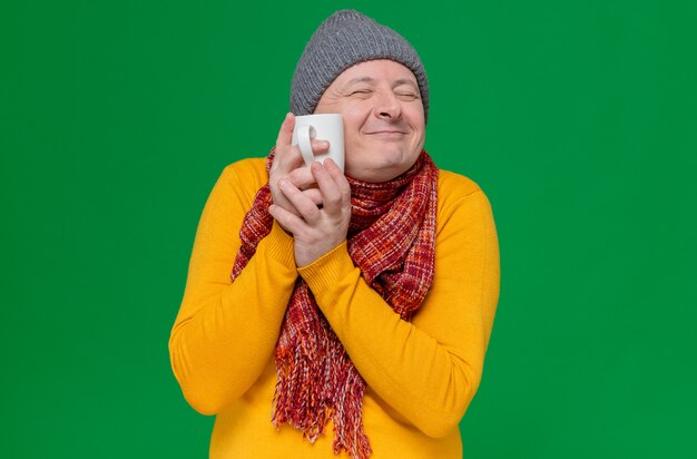 Pleased adult slavic man with winter hat and scarf around his neck holding cup close to his face