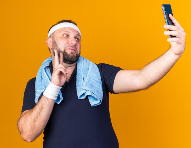 довольный взрослый славянский спортивный мужчина с полотенцем на шее, с повязкой на голову и браслетами, делающий селфи, жестикулирующий со знаком победы, изолированным на оранжевой стене с копией пространства
