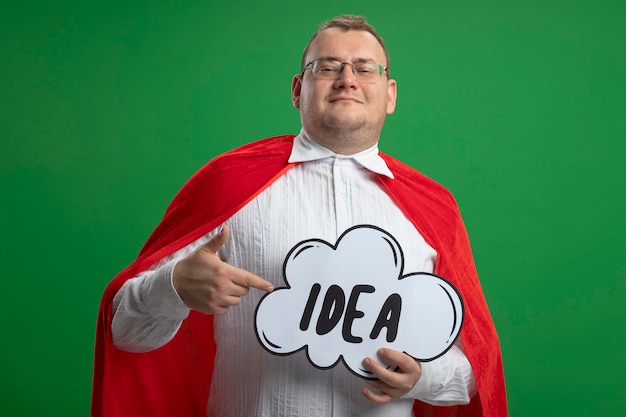 Free photo pleased adult slavic superhero man in red cape wearing glasses holding and pointing at idea bubble  isolated on green wall