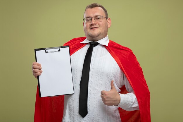 Pleased adult slavic superhero man in red cape wearing glasses and tie holding clipboard looking at camera showing thumb up isolated on olive green background