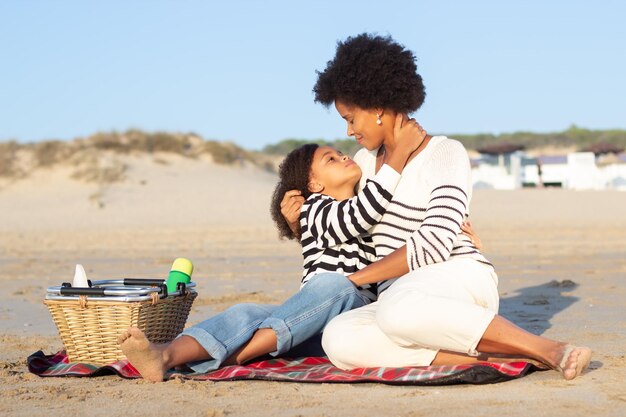 Довольная афроамериканская семья на пикнике на пляже. Мать и дочь в повседневной одежде сидят на одеяле, обнимаются. Семья, отдых, концепция природы