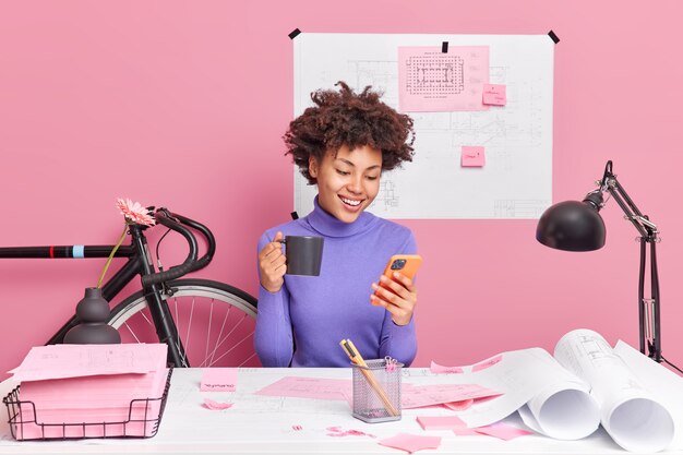 Pleased African American woman using smartphone, drinks coffee, dressed in casual jumper poses at desktop with papers around works on future engineer project