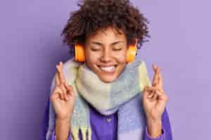 Бесплатное фото Довольная афро-американская девочка-подросток держит глаза закрытыми, кусает губы суеверно скрещивает пальцы на удаче, носит наушники на ушах, шарф на шее