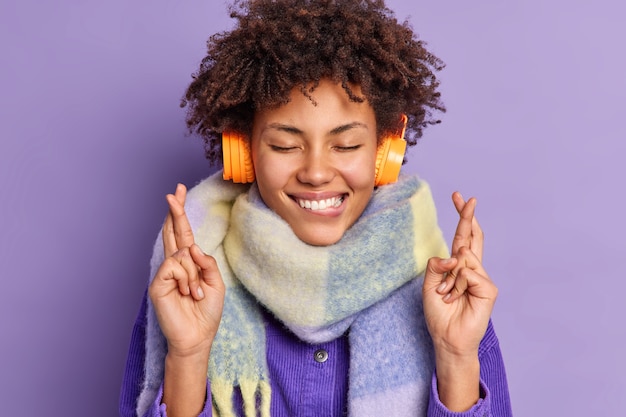 Pleased Afro American teenage girl keeps eyes closed bites lips stands superstitious crosses fingers for good luck wears headphones on ears scarf around neck