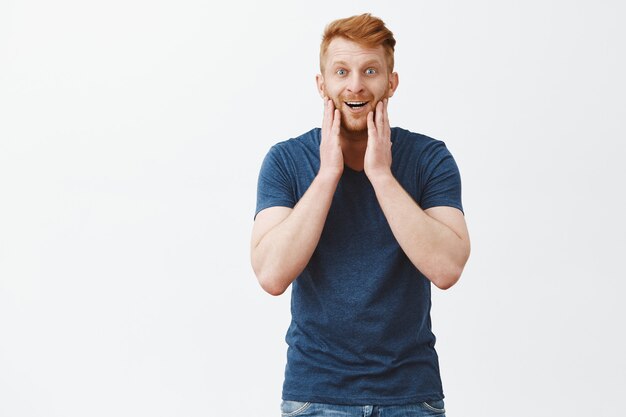 Pleased and amazed guy looking in mirror, satisfied with new haircut. Good-looking masculine and handsome redhead male model in blue t-shirt, touching cheeks and gazing with broad smile