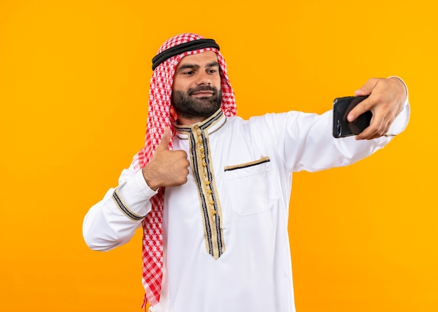 オレンジ色の壁の上に立って自信を持って見える彼のスマートフォンを使用して自分撮りを取っている伝統的な服を着て喜んでアラビアのビジネスマン