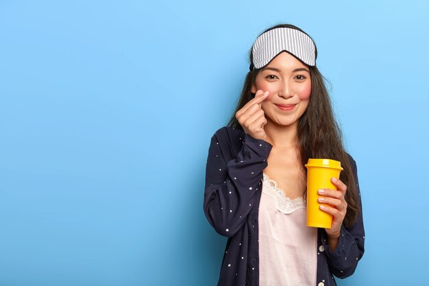 Pleased Asain dark haired woman makes korean like gesture, dressed in pyjamas and sleep mask, holds yellow takeout coffee cup