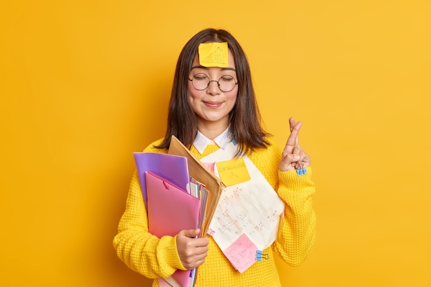 Pleased Asain female student believes in good luck at exam stands with eyes closed and fingers crossed believes dreams come true stuck with papers holds folders.