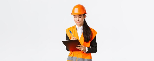 Pleased asian female construction engineer architect taking notes in clipboard writing down something during inspection at building area wearing safety helmet businesswoman inspect workers