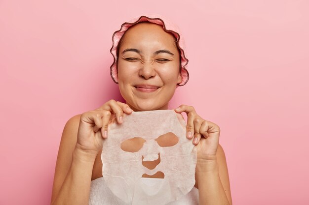 Pleased Asian female holds facial sheet mask, going to apply on face, gets pleasure from beauty treatments, keeps eyes closed