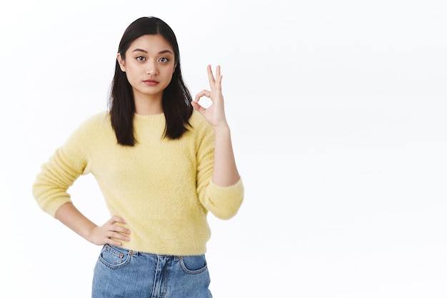 Pleased asian female team lead saying not bad to employee Satisfied confident young woman in yellow sweater showing okay approval or agree gesture smiling pleased good choice white background