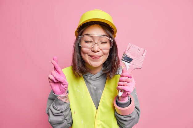 Pleased Asian woman crosses fingers holds paint brush renovates house makes wish believes in good luck wears safety glasses helmet gloves