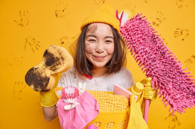 Pleased Asian woman gets pleasure from cleaning process holds dirty sponge and mop poses near basket full of laundry isolated over yellow wall