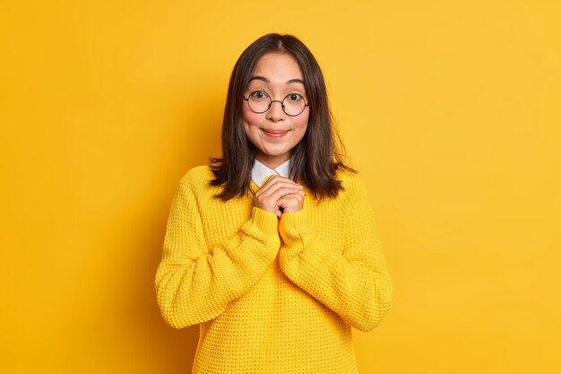 Pleased Asian woman keeps hands together anticipates for something good smiles gently has dimples on cheeks dressed in casual clothes.